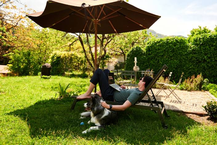 house sitter reclining under an umbrella while working on a laptop patting a black and white dog next to them in the sunshine